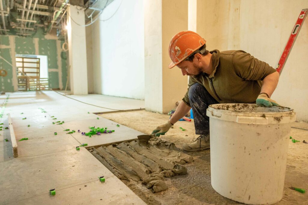 A construction worker lays tiles in an indoor renovation project, wearing safety gear.